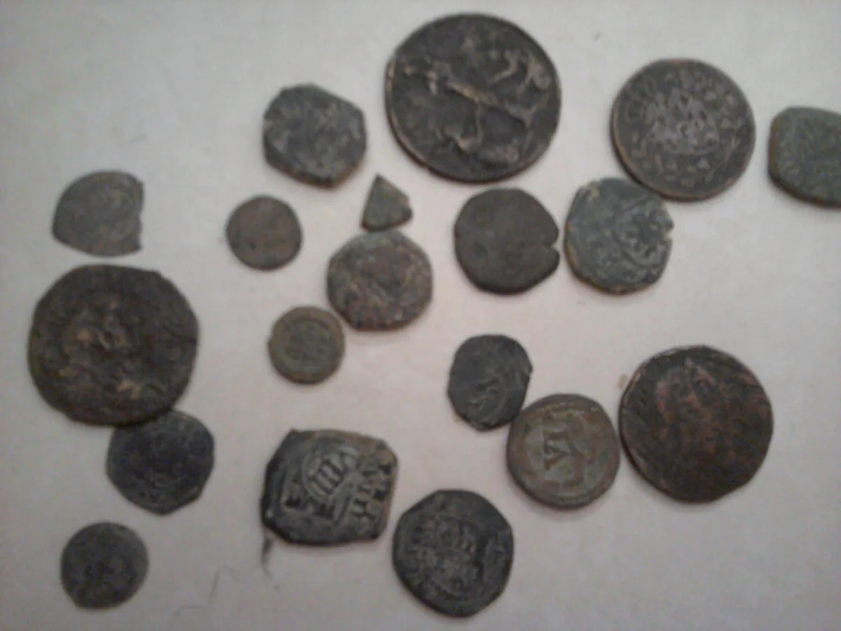 A collection of old, corroded, and irregularly shaped coins and fragments lie scattered on a plain white surface. The…
