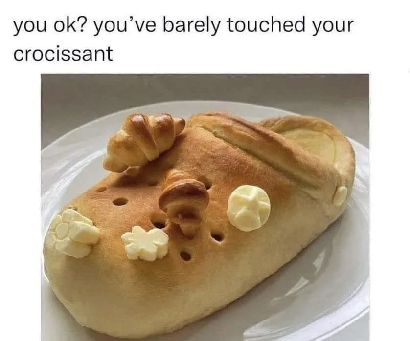 A baked good shaped like a Croc shoe sits on a white plate, decorated with small croissant pieces and butter pats resembling…