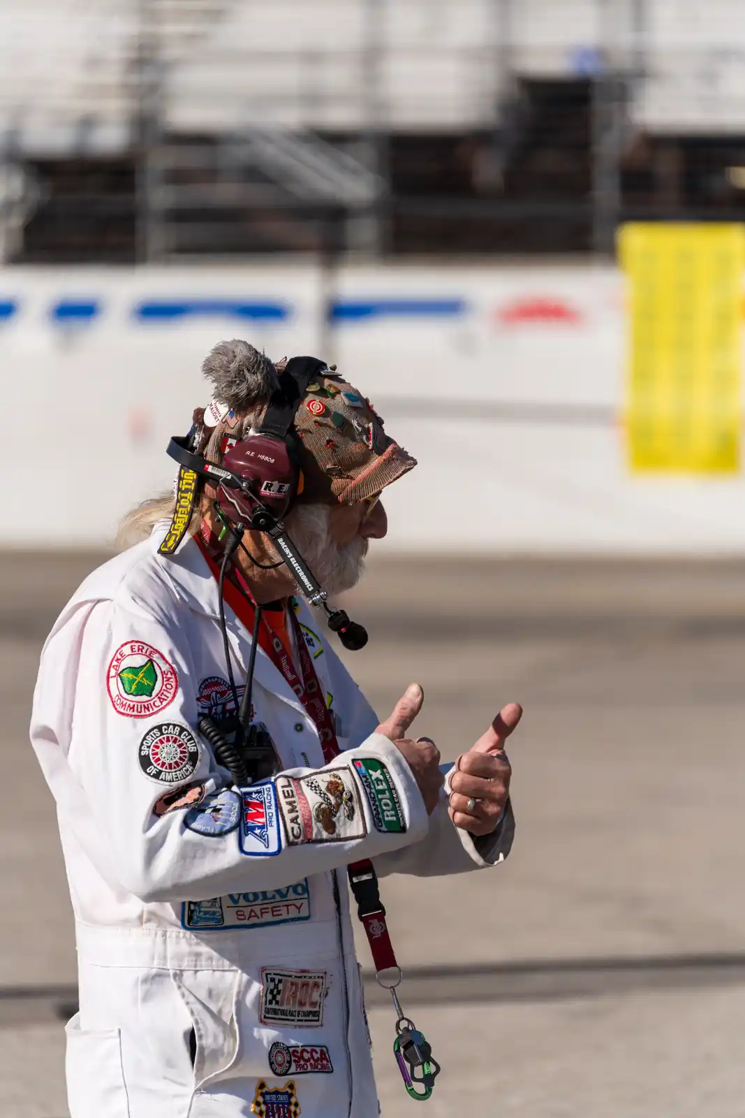 An older man in a white racing jacket covered in patches and wearing a headset and a heavily decorated cap gives two thumbs…