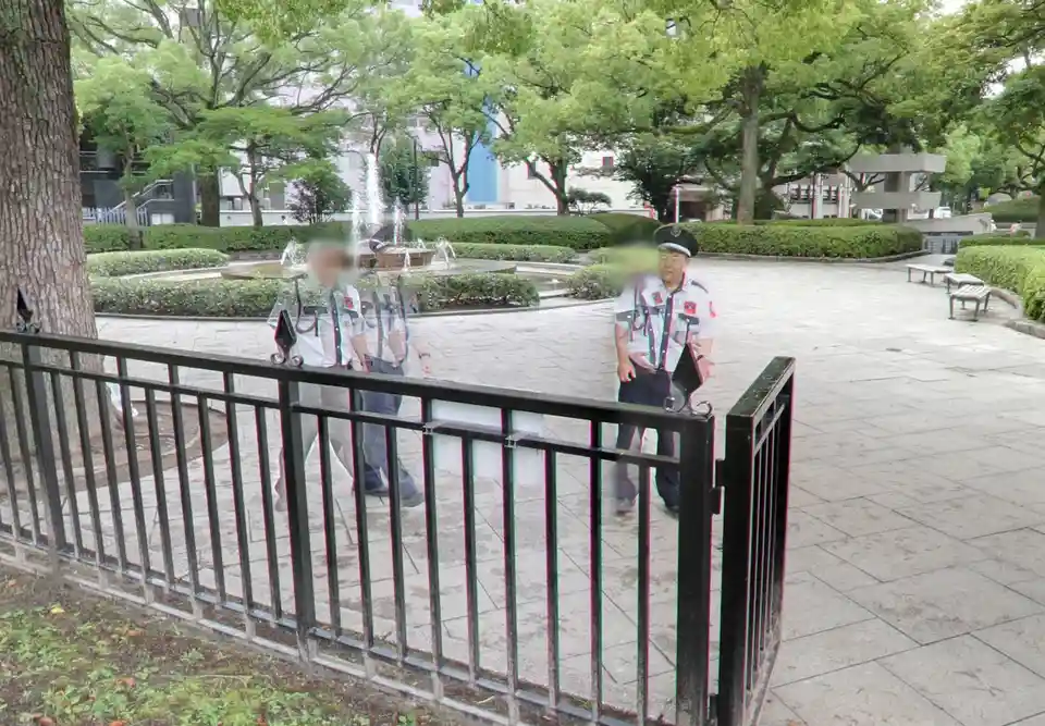 Two uniformed Japanese park guards stand behind a black metal fence in a park with trees, benches, and a fountain in the…