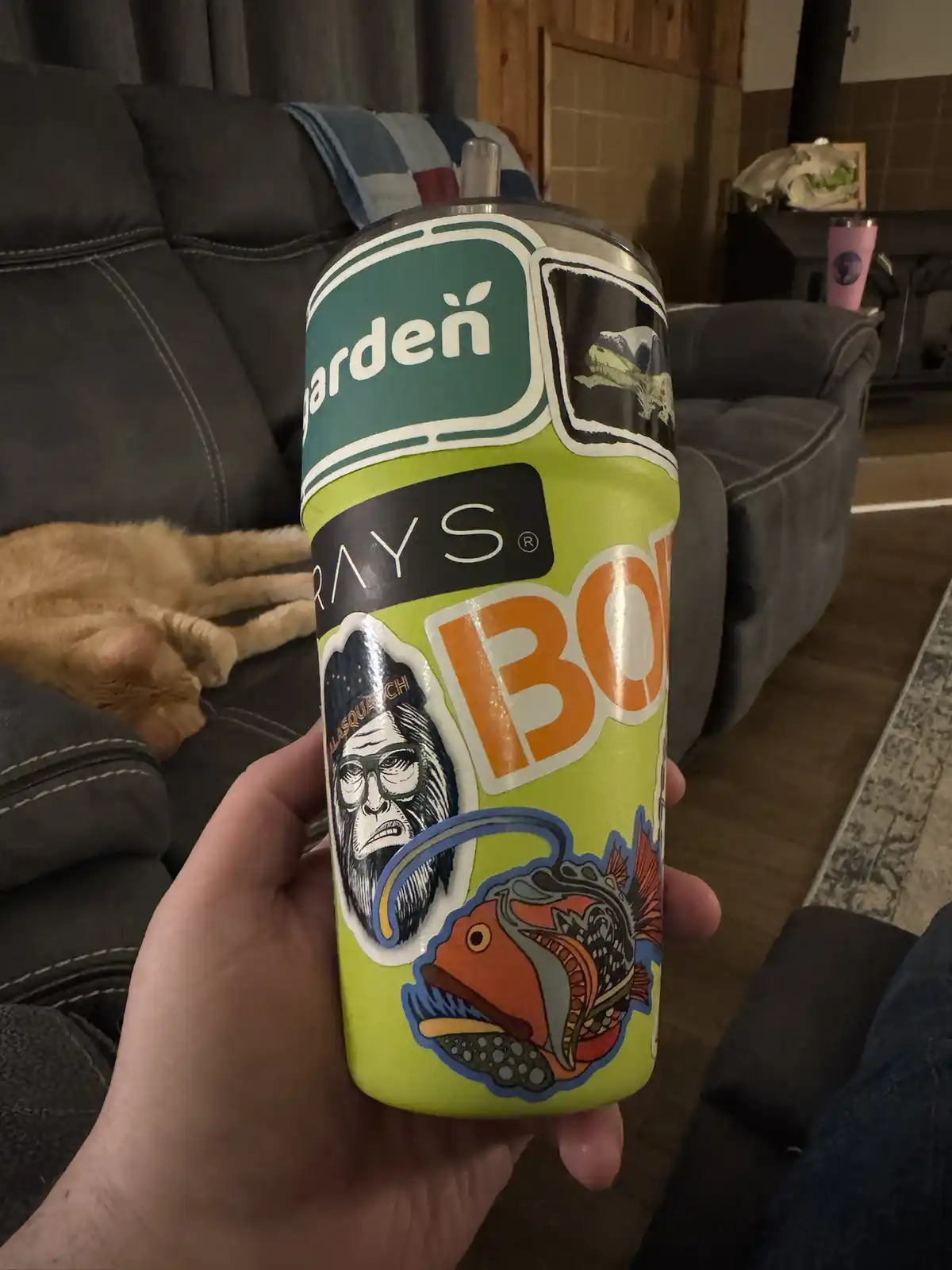 A hand holds a lime-green tumbler decorated with stickers, including a 'Garden Rays BO' label, a Sasquatch, and a fish. A…