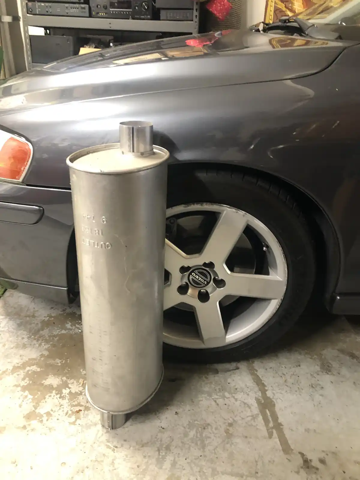 A large, cylindrical, silver metal exhaust pipe stands upright on a concrete garage floor next to the front wheel of a dark…