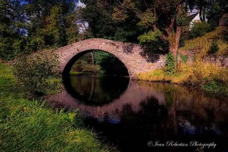 A picturesque stone arch bridge spans a calm, reflective river surrounded by lush greenery and trees under a partly cloudy…