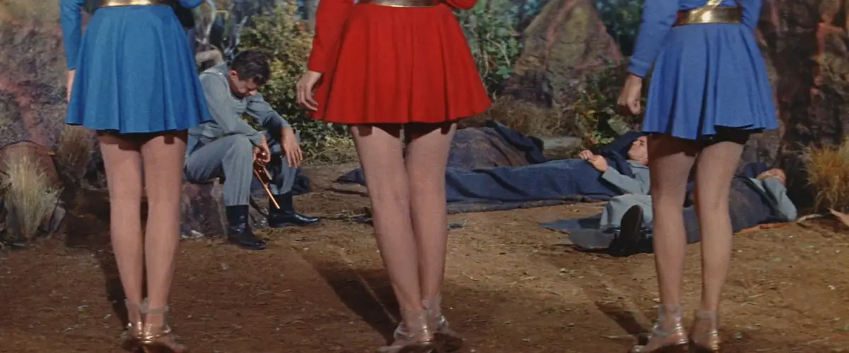 Three women in colorful, short skirts and gold belts stand with their backs to the camera, obscuring the view of two men in…