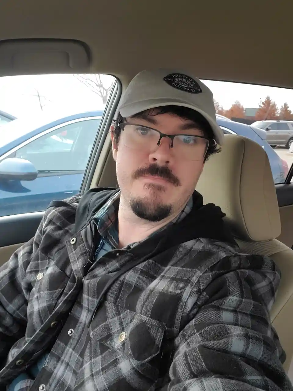 A man with glasses and a mustache sits in the driver's seat of a car, wearing a plaid shirt and a baseball cap. He stares…