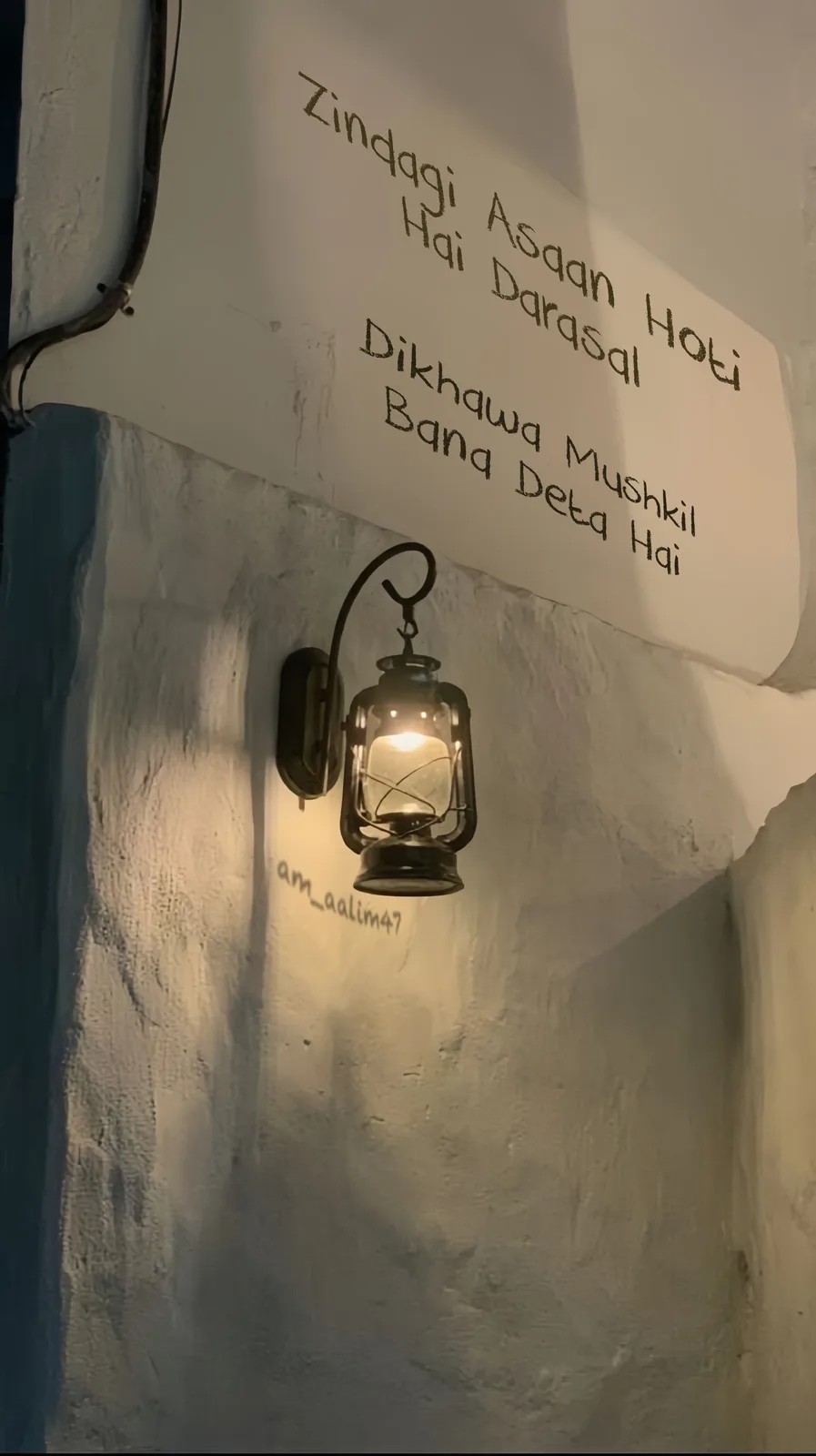 A warmly lit vintage lantern hangs on a textured white wall, casting a soft glow. Above it, handwritten Hindi text reads…