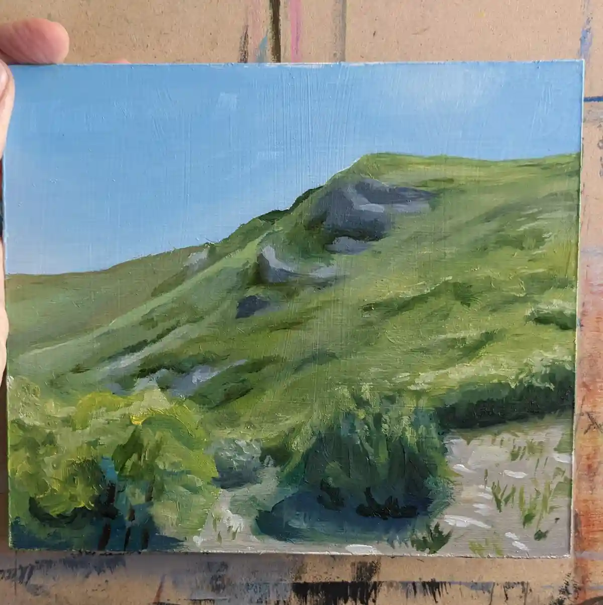 A hand holds up a small, textured oil painting depicting a green, rolling hill under a clear blue sky, with brushstrokes…