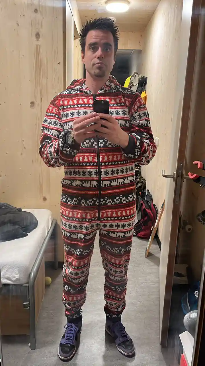 A man stands in a narrow wooden room, taking a mirror selfie while wearing a full-body, red, black, and white patterned…