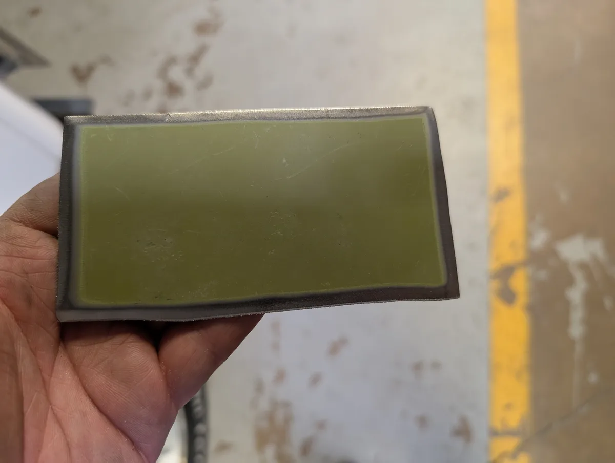 A hand holds a rectangular, olive-green, translucent glass or plastic panel with visible scratches and smudges, framed by a…