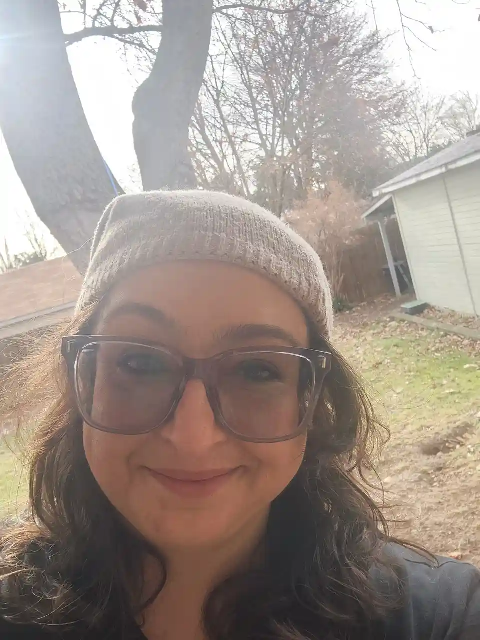 A selfie of a woman with dark curly hair, wearing a beige knit beanie and large brown-framed glasses, smiling slightly while…