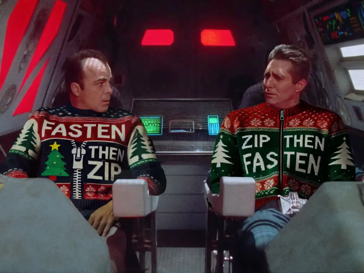 Two men in a spaceship cockpit wear Christmas sweaters with zippers; one says 'FASTEN THEN ZIP' and the other 'ZIP THEN…