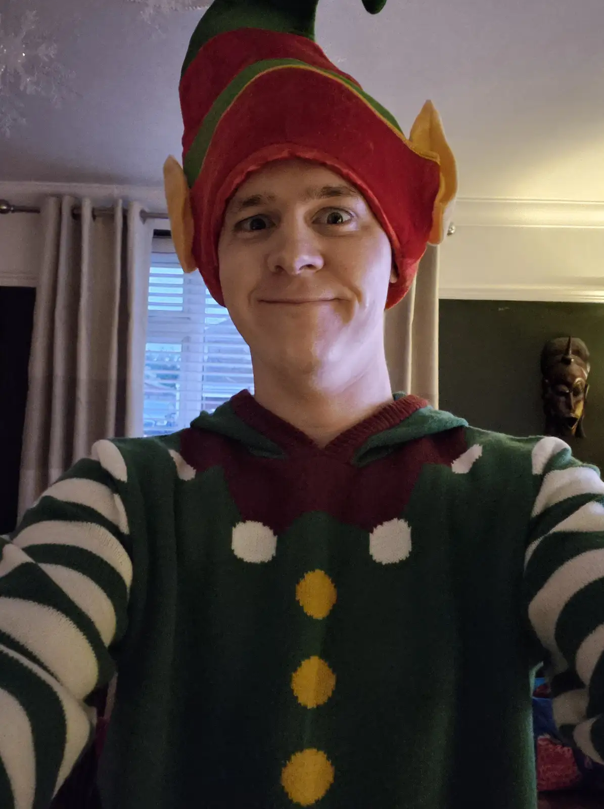 A person wearing a red and green elf hat with yellow ears and a matching green, red, and white striped elf sweater takes a…