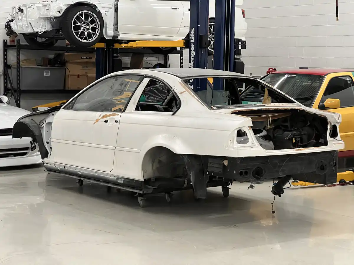 A white car body, stripped of its interior and most panels, sits on wheels in a garage, with other cars and lifts visible in…