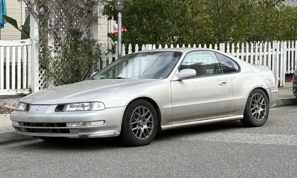 A silver Honda Accord coupe is parked on a residential street in front of a white picket fence, covered in a thick layer of…