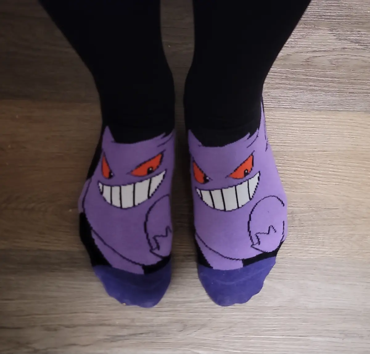 A close-up shot shows a person's feet wearing purple socks featuring the cartoon character Gengar from Pokémon, which has a…