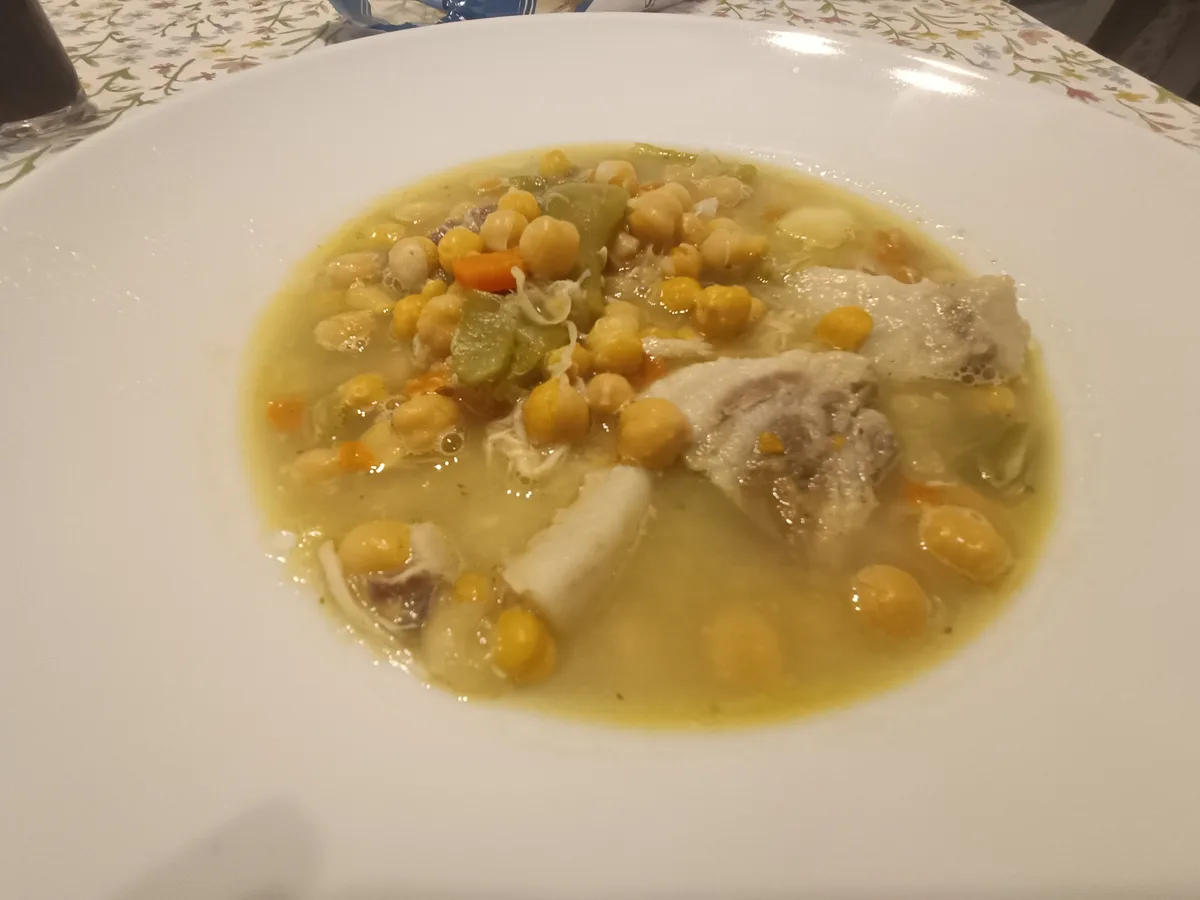 A close-up photo shows a white bowl filled with a yellowish broth containing chickpeas, small pieces of meat, and vegetables…