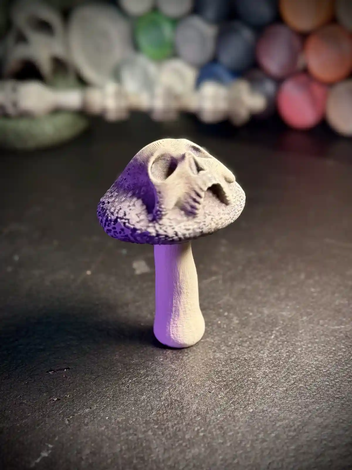 A small, hand-painted mushroom figurine with a purple cap and stem, featuring a carved human-like face on its cap. The…