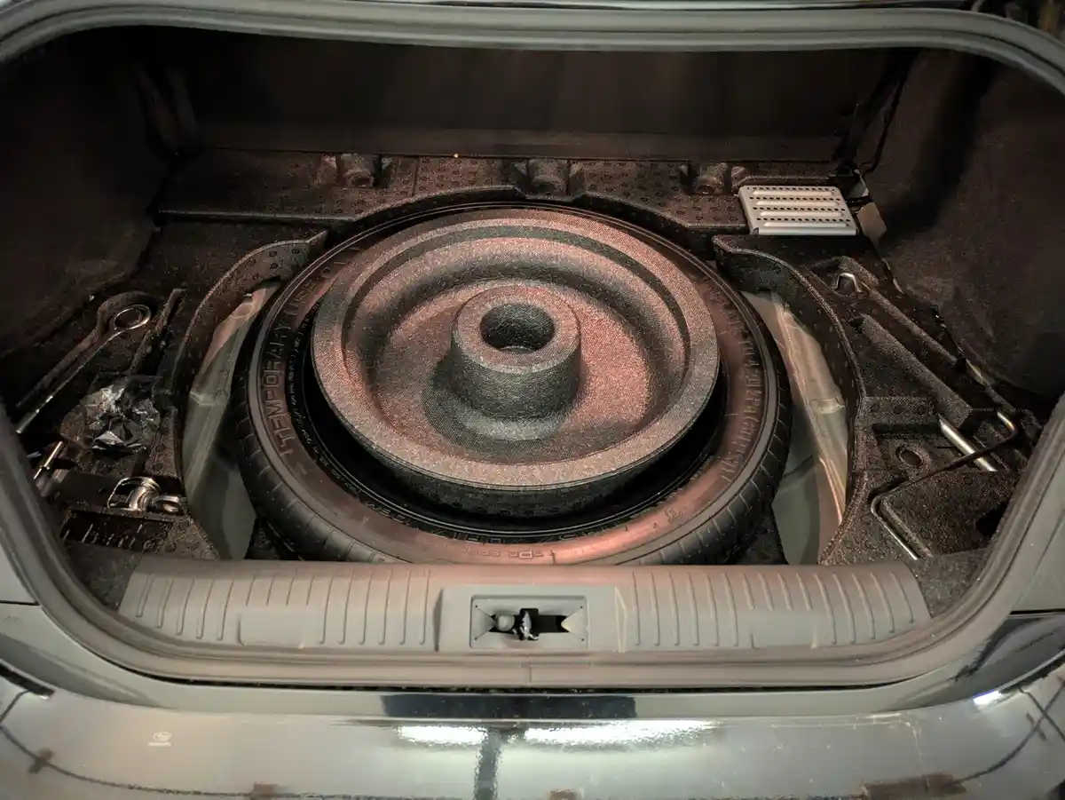 The image shows the open trunk of a car, revealing a spare tire mounted in its designated space. The tire has visible text…