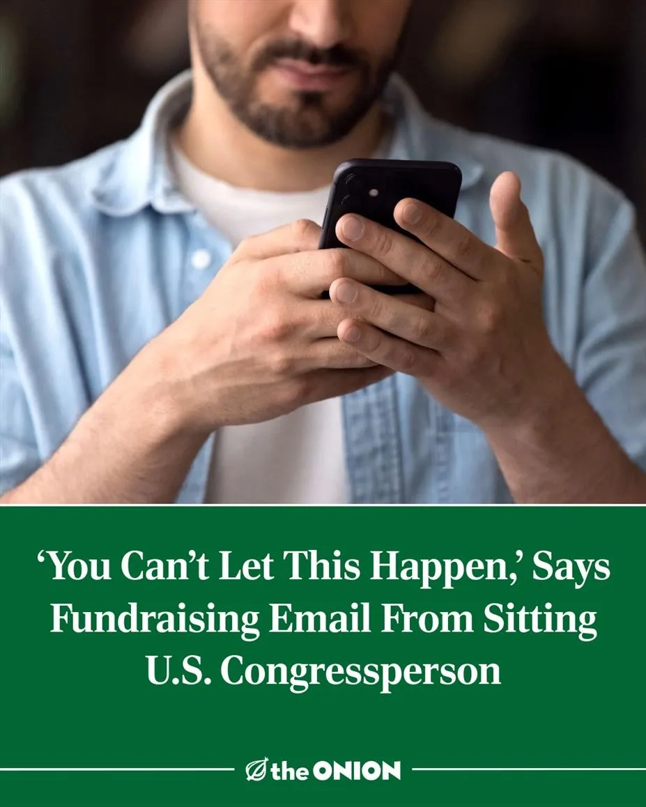 A man with a beard wearing a light blue shirt and white undershirt is looking down at a black smartphone held in both hands…