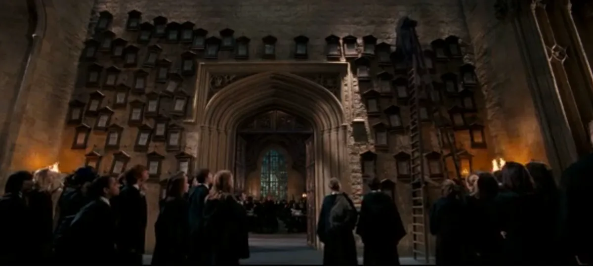 A wide shot from the Harry Potter movies shows students in dark robes standing in a large, stone-walled hall with many…
