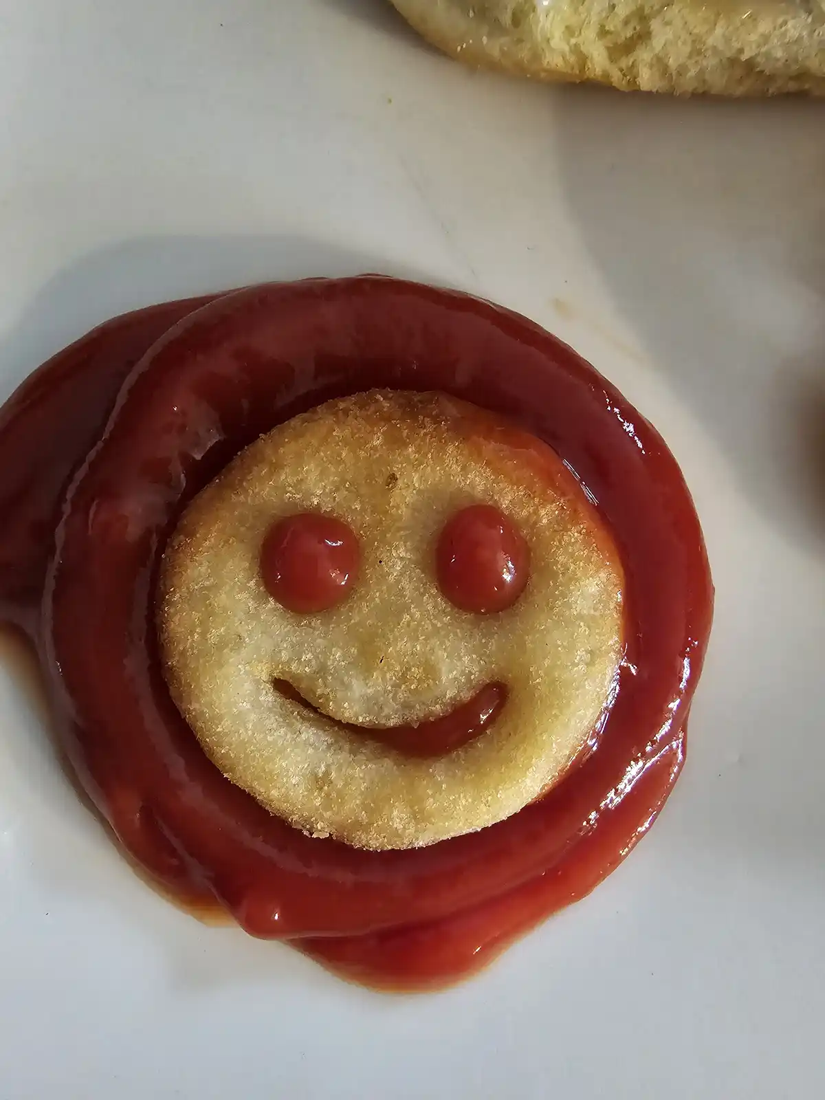 A close-up of a fried food item, likely a hash brown or onion ring, arranged to resemble a smiling face with ketchup. Two…