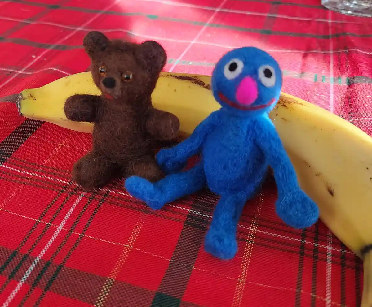 Two felt toys, a brown teddy bear and a blue Grover-like figure, sit on a red plaid tablecloth next to a yellow banana. The…