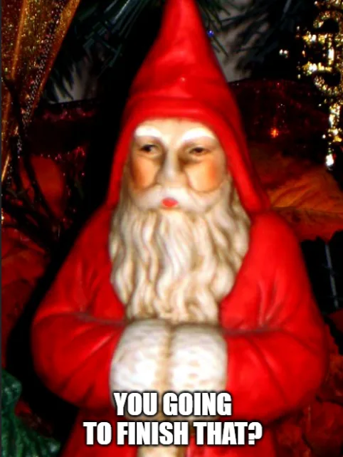 A plastic Santa Claus figurine with a long white beard and red suit stands with arms crossed, looking sternly at the viewer…