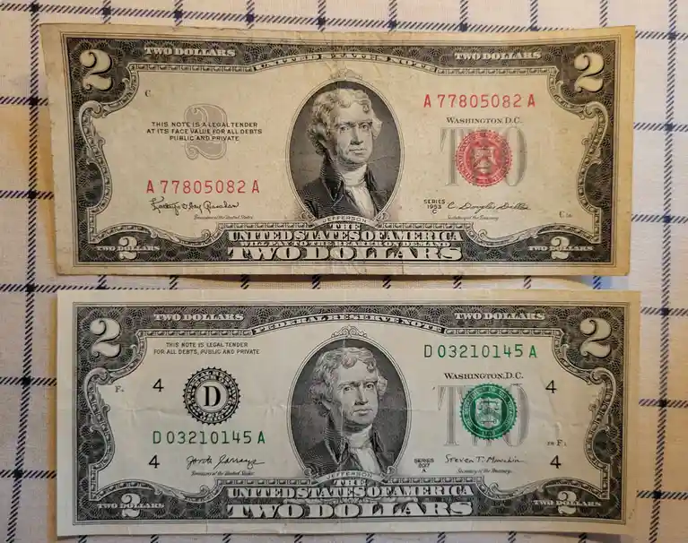 Two U.S. two-dollar bills are laid out side-by-side on a plaid surface, one from 1953 with a red seal and one from 2017 with…