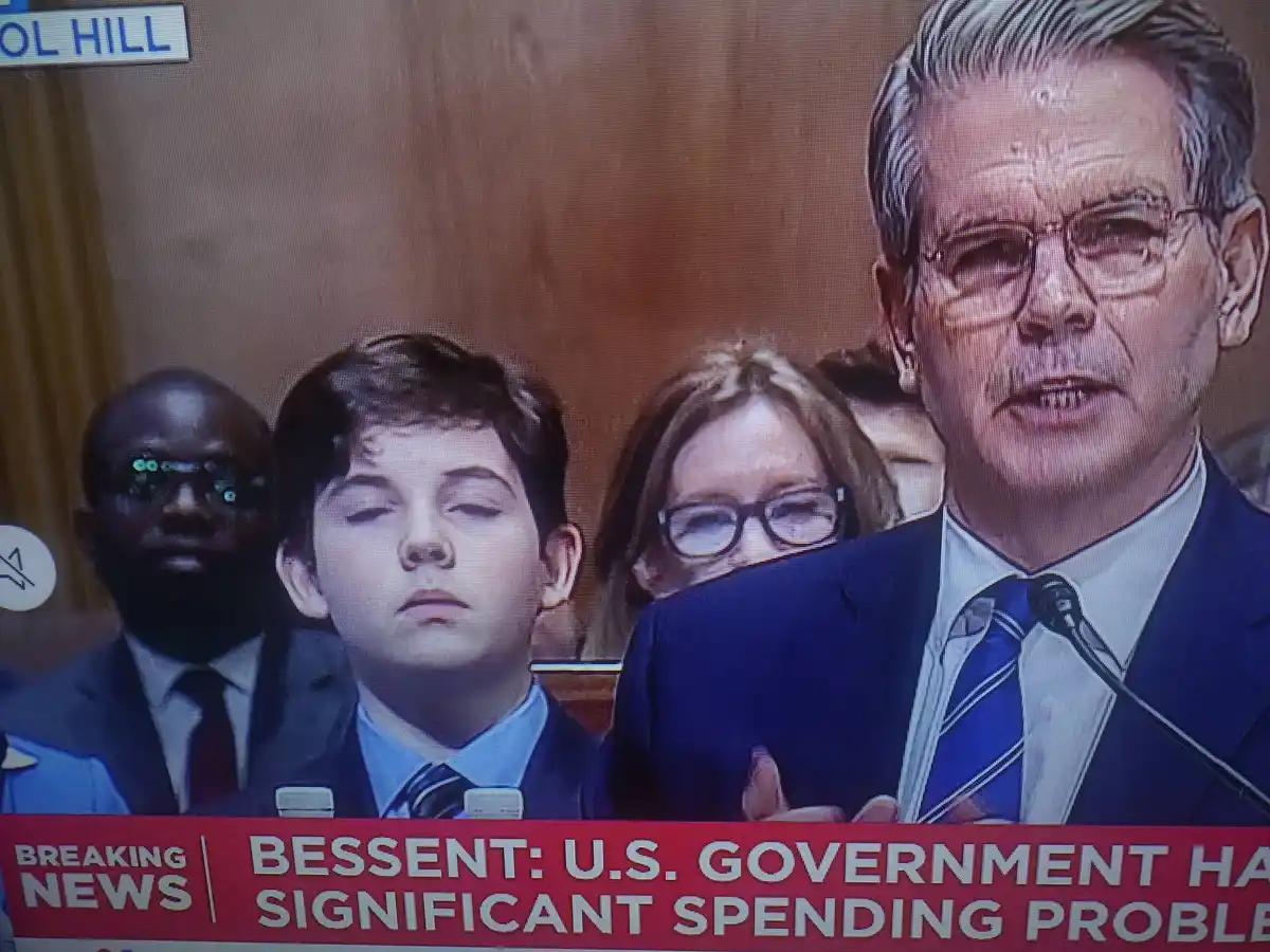 A screenshot of a televised hearing shows a man speaking at a podium while three people sit beside him. The person in the…