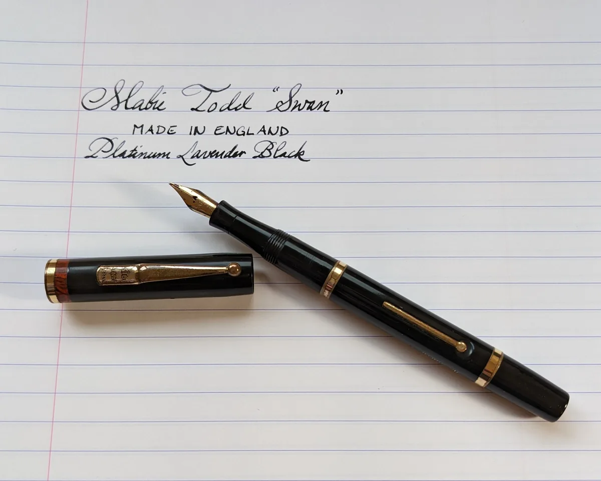 A black fountain pen with gold trim and a wooden cap band lies on lined notebook paper next to its cap. Above the pen…