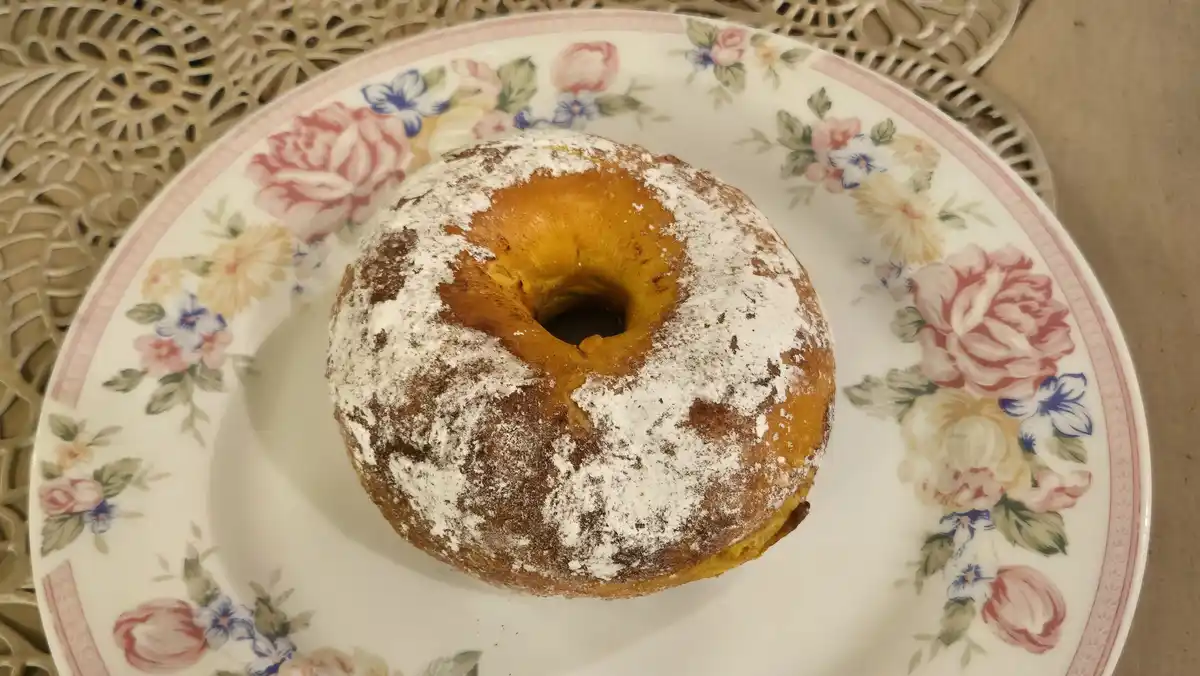 A single, powdered sugar-dusted donut sits centered on a white ceramic plate decorated with a delicate floral pattern of…