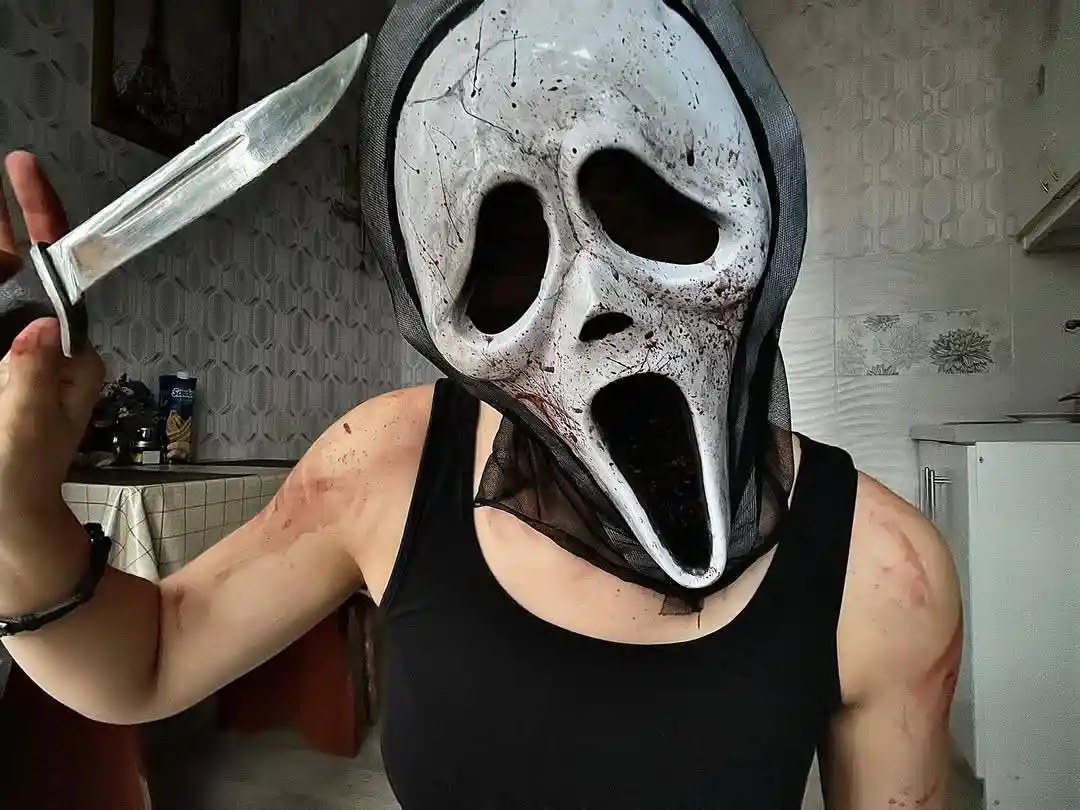A person wearing a white, blood-splattered Ghostface mask from the 'Scream' movie franchise, holding a large kitchen knife…