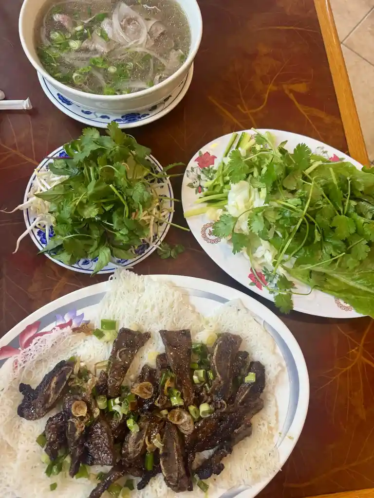 A table displays a meal with a bowl of pho soup, plates of fresh herbs and bean sprouts, and a plate of grilled beef ribs…