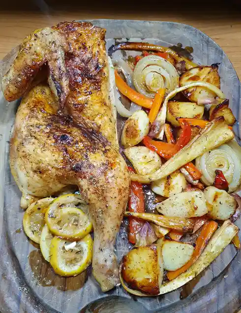 A roasted whole chicken with golden-brown skin sits on a wooden platter next to roasted vegetables including onions…
