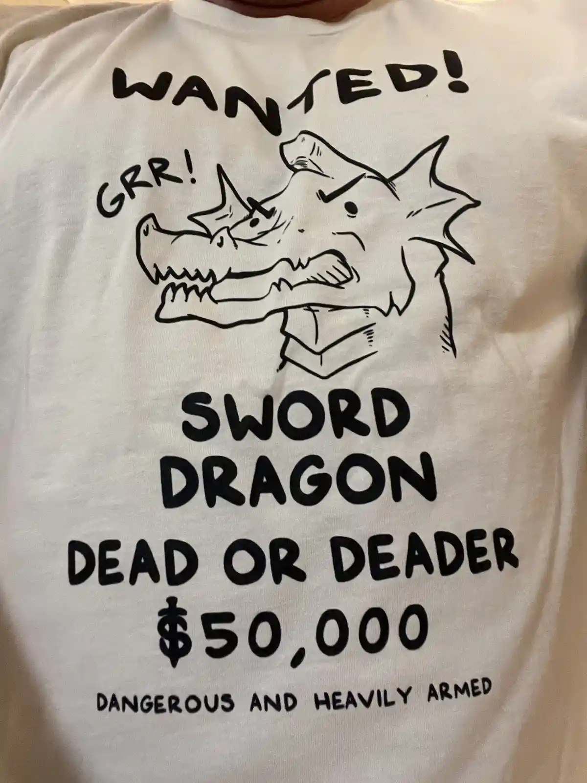 A white t-shirt displays a black ink drawing of a cartoon dragon with a sword in its mouth, roaring 'GRR!'. The text mimics…