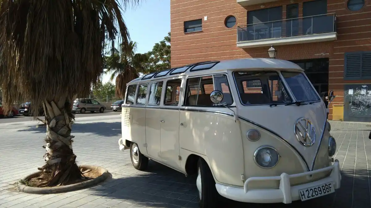 A cream-colored vintage Volkswagen Type 2 van with a roof rack is parked on a paved area next to a palm tree. The van has a…