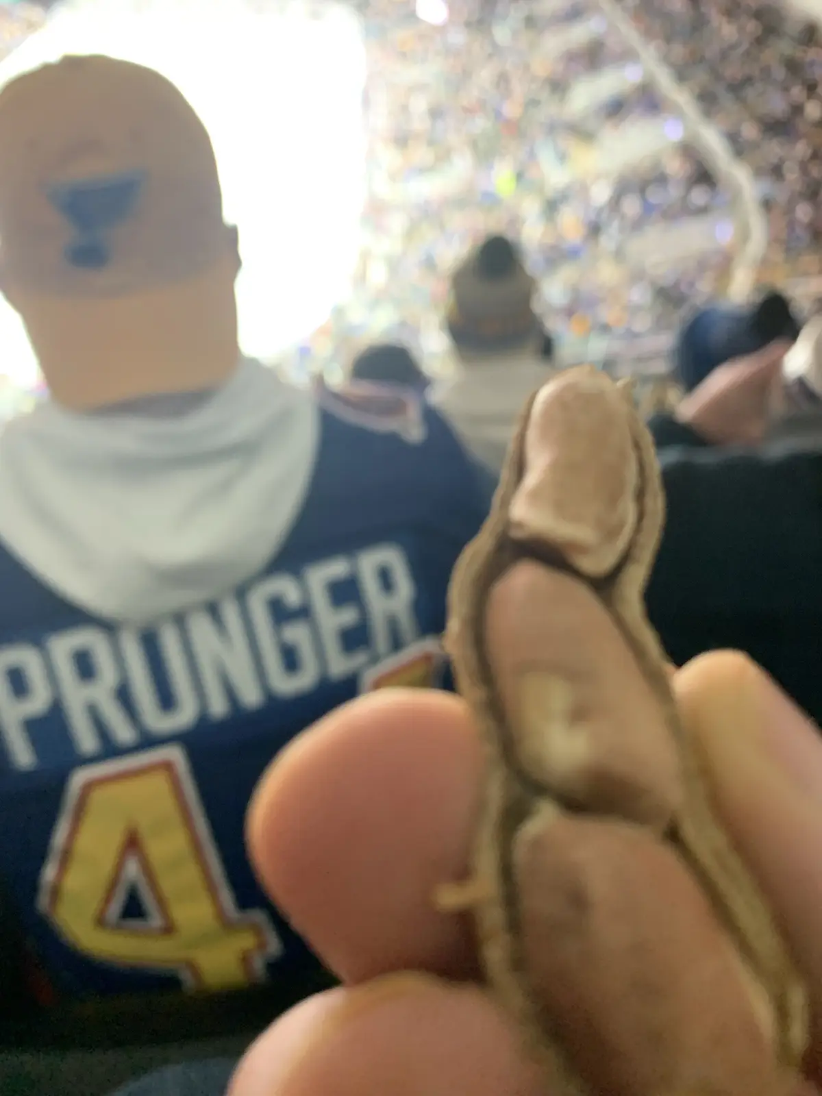 A close-up shot shows a hand holding a single peanut in its shell, with a blurred background of a crowded sports arena. In…