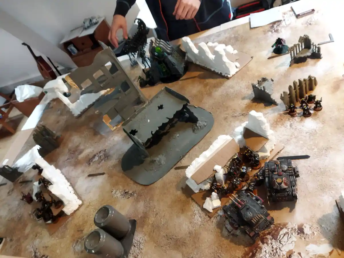 An overhead shot of a tabletop wargaming setup with miniature figures, terrain pieces, and a person's hands adjusting a…