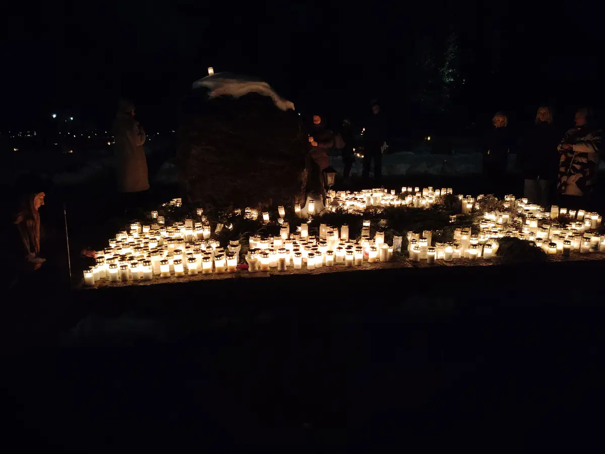 A nighttime photo shows a large gathering of people around a massive stone monument covered in snow, surrounded by hundreds…
