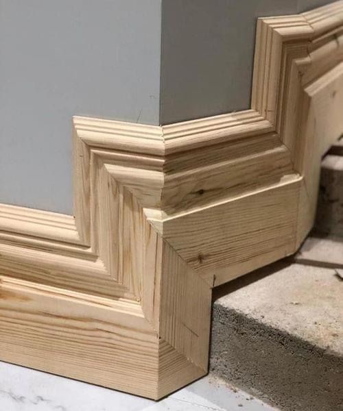 A close-up photo shows an ornate, unfinished wooden baseboard molding at a wall corner, where the two pieces are visibly…