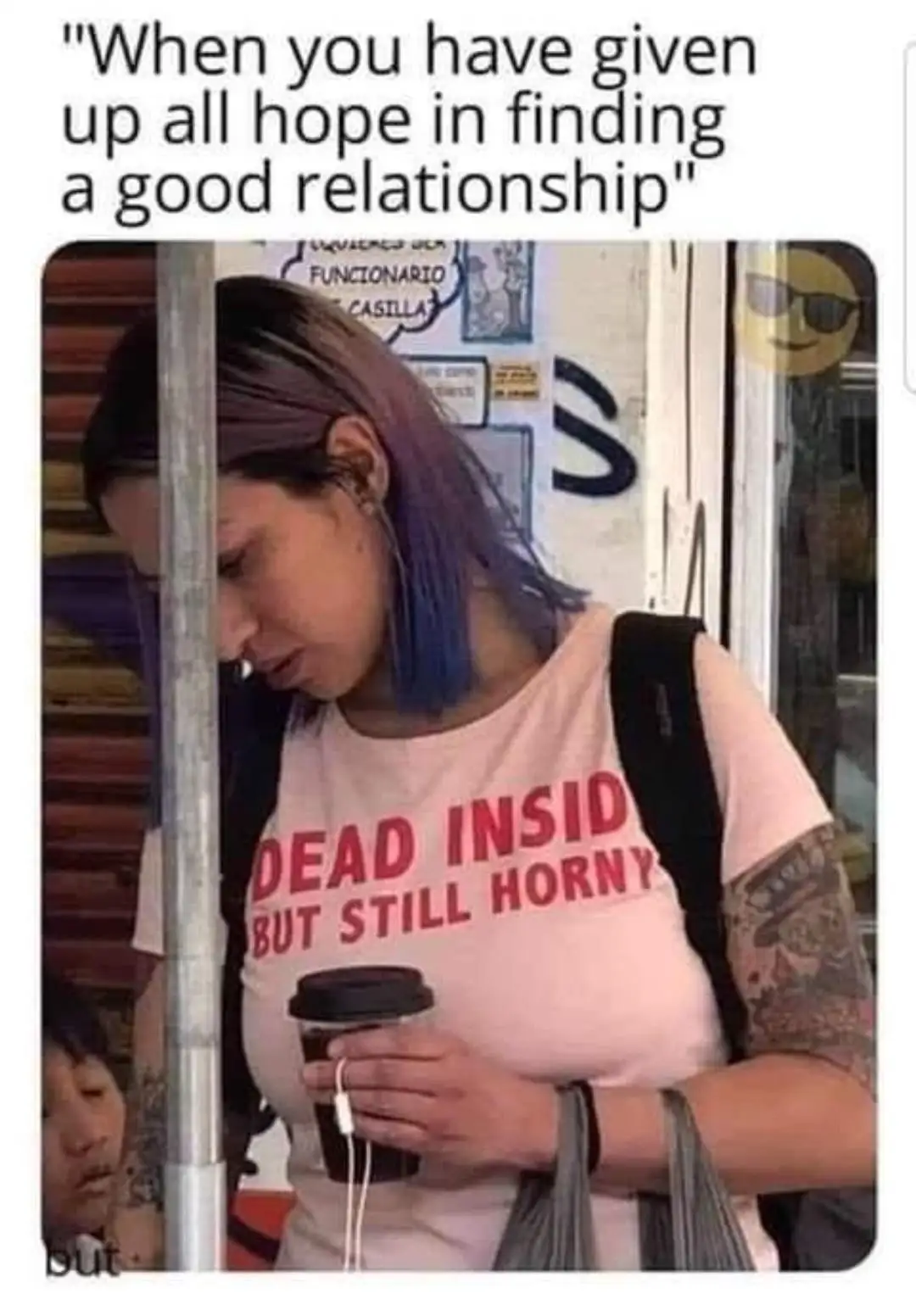 A woman with purple-blue hair and tattoos on her arms, wearing a pink t-shirt that reads 'DEAD INSIDE BUT STILL HORN', holds…