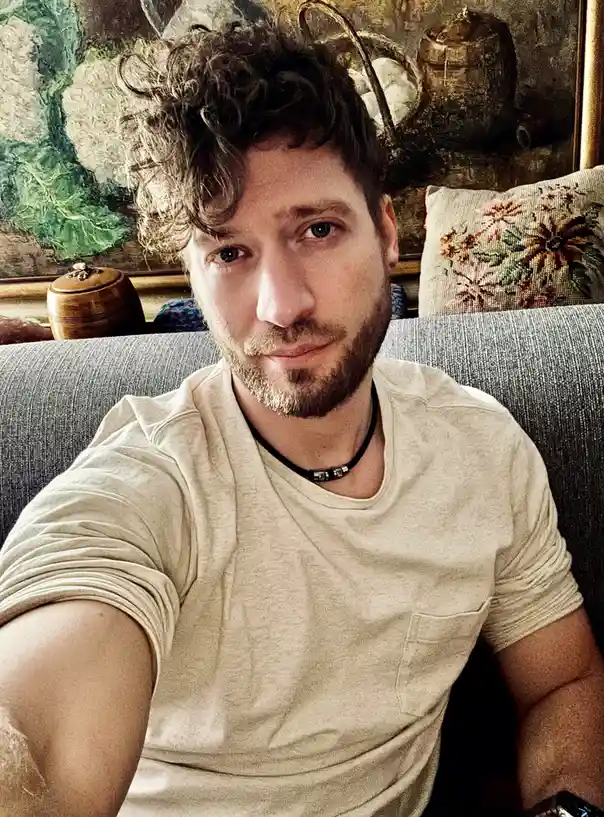 A man with curly hair and a beard sits on a gray couch, wearing a light-colored t-shirt and a black beaded necklace, looking…