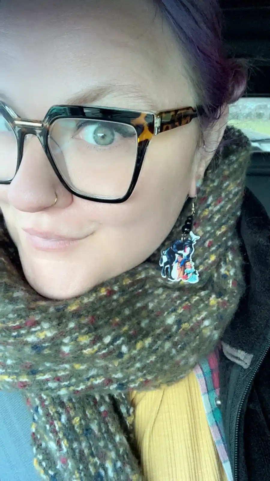 A close-up selfie of a person with purple hair, wearing tortoiseshell glasses and a colorful scarf, showing a small earring…