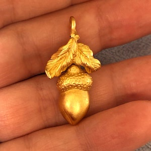 A close-up photo shows a hand holding a small, shiny gold acorn-shaped pendant with detailed leaves. The acorn is resting on…
