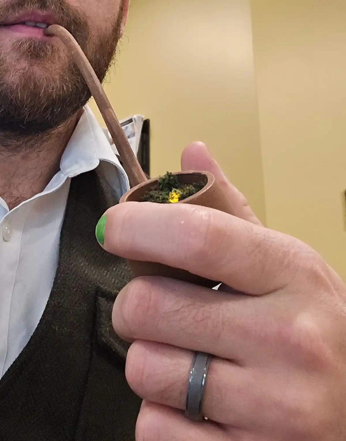 A close-up photo shows a person with a beard smoking a small wooden pipe, holding it with their right hand. The person is…