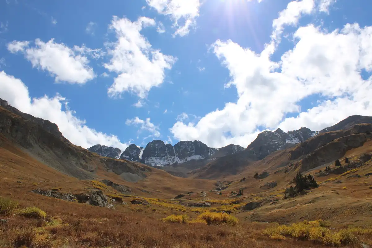 A scenic mountain landscape with rugged peaks, snow-capped ridges, and golden-brown autumnal vegetation under a bright blue…