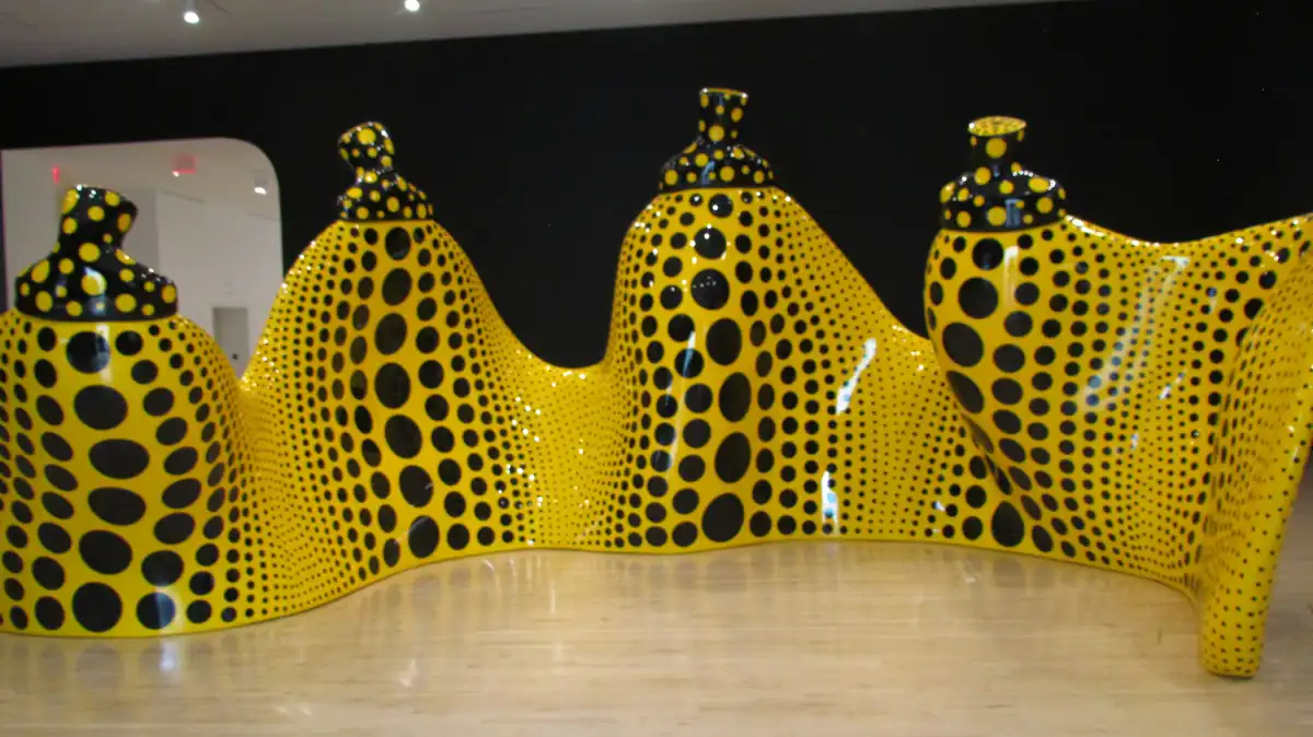 a large, wavy, yellow sculpture covered in black polka dots, resembling a bench but with a surreal, artistic design, sits on…