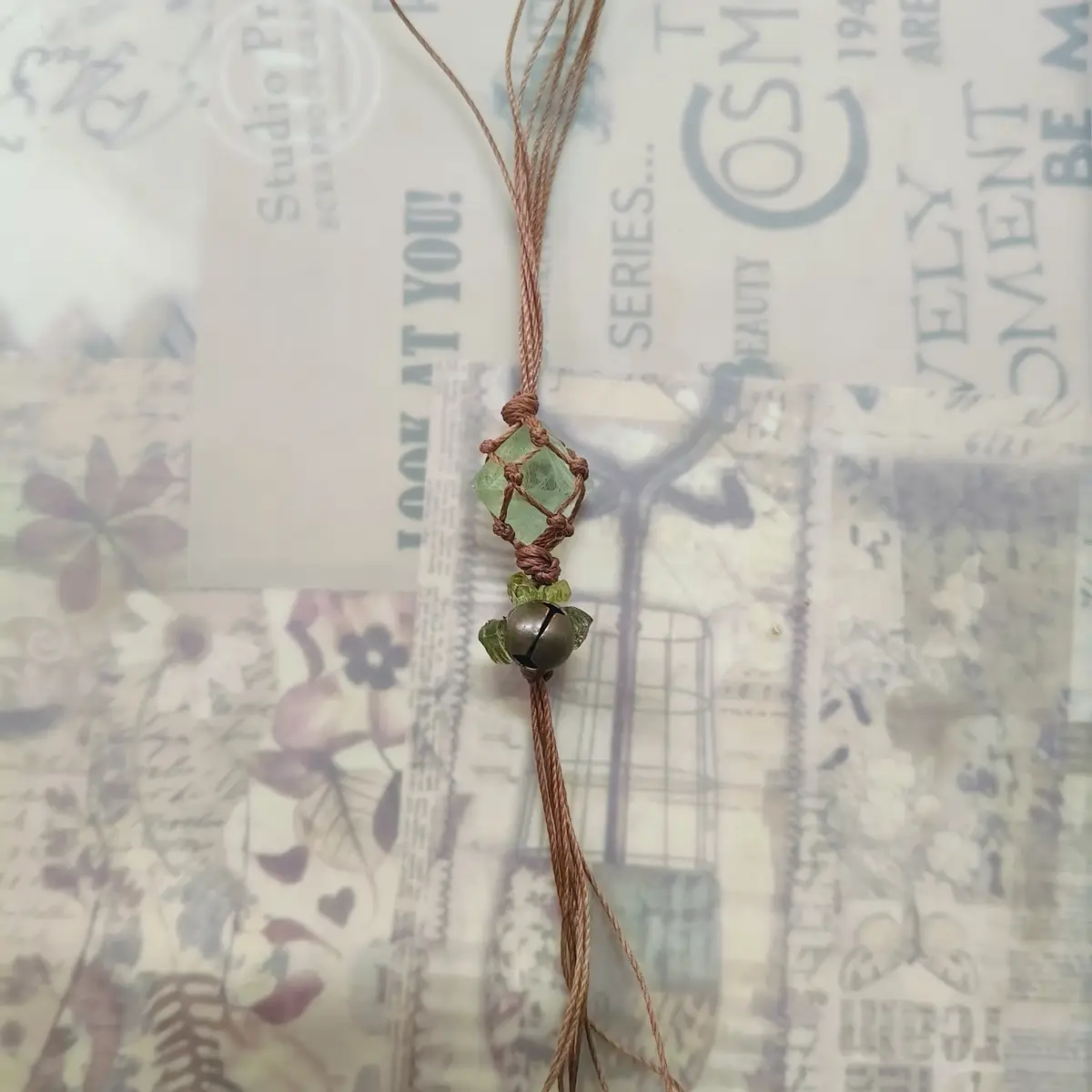 A handmade macrame pendant with a green gemstone and a small bell, strung on brown cord, rests on a collage of vintage-style…
