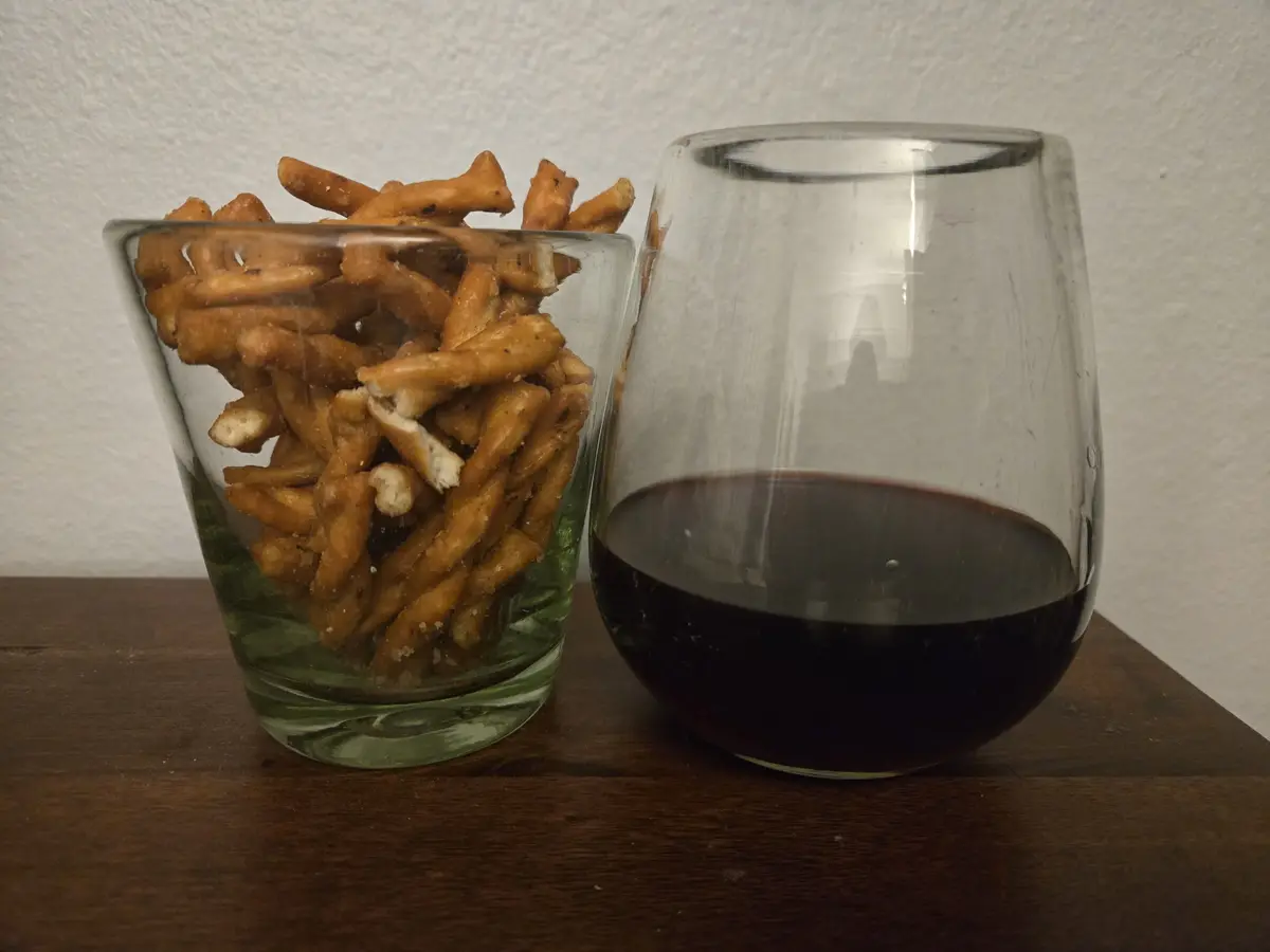 Two glasses sit side by side on a wooden surface: one filled with golden-brown pretzels and the other with dark red wine…
