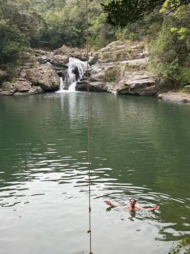 A man is swimming in a green pool below a waterfall, smiling and gesturing with open arms. A thick rope hangs vertically…